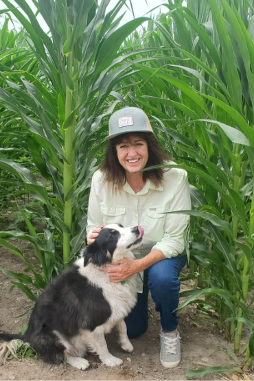 Ann kneeling with her dog with a corn field background